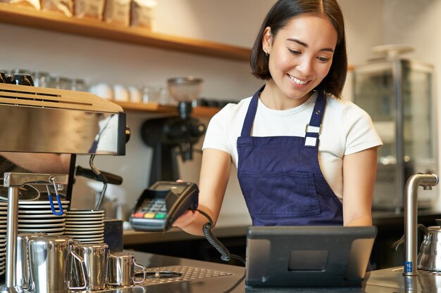 コーヒー ショップのカウンターで働く POS 端末で注文を処理する笑顔のアジアのバリスタの女の子