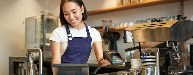 Free photo smiling asian barista girl processing order enter order in pos terminal working at counter in coffee