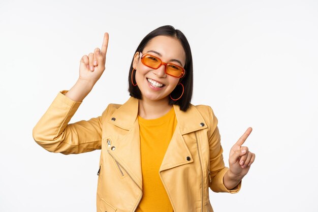 Smiling asian brunette woman in sunglasses pointing fingers sideways left and right showing variants laughing and dancing wearing sunglasses white background