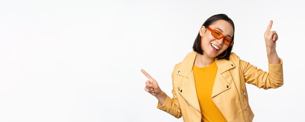 Улыбающаяся азиатская брюнетка в солнцезащитных очках, указывающая пальцами влево и вправо, показывая варианты смеха и танца в солнцезащитных очках на белом фоне