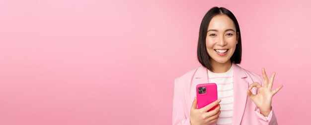 Улыбающаяся азиатская деловая женщина показывает знак "хорошо" при использовании приложения для мобильного телефона, рекомендуя приложение для смартфона, стоящее на розовом фоне