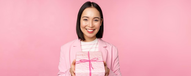 Smiling asian businesswoman in suit giving you gift in wrapped box standing over pink background