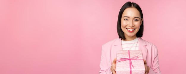 분홍색 배경 위에 서 있는 포장된 상자에 선물을 주는 정장을 입은 웃고 있는 아시아 여성