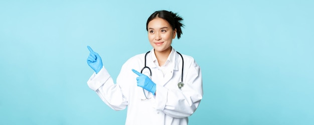 Sorridente medico asiatico lavoratore medico in uniforme che punta e guardando a sinistra lo spazio della copia in piedi su sfondo blu