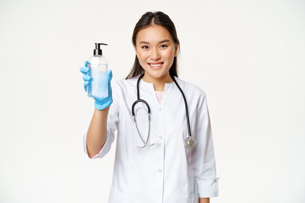 Smiling asian doctor showing hand sanitizer bottle of antiseptic wearing sterile rubber gloves and c...