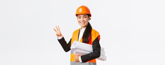 Smiling asian female architect at construction area building estates showing number three carry blueprint as wearing safety helmet giving fixed time for task done white background