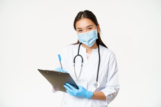 Smiling asian female doctor in face mask and gloves writing down patient diagnose prescription holdi...