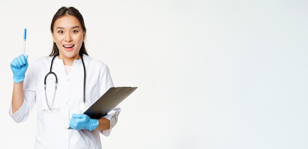 Free photo smiling asian female doctor raising pen up eureka gesture holding clipboard standing in medical unif