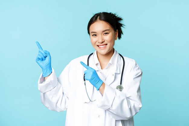 웃고 있는 아시아 여성 의사, 치료사는 왼쪽 상단 모서리를 손가락으로 가리키며 의료 광고를 보여주고 파란색 배경 위에 서 있습니다.