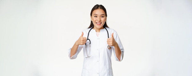 Smiling asian female doctor wearing medical robe showing thumbs up pleased recommending health produ