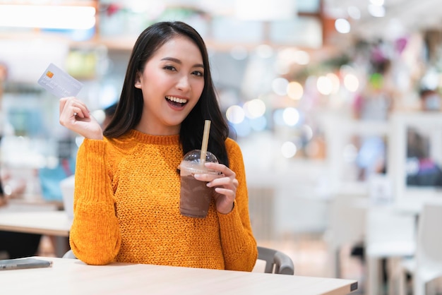 Улыбающаяся азиатская женщина держит за руку напиток со льдом, сидит, расслабляется в кафетерии, фуд-корт, универмаг маллазийская женщина, держит кредитную карту, концепция безналичного образа жизни, покупки