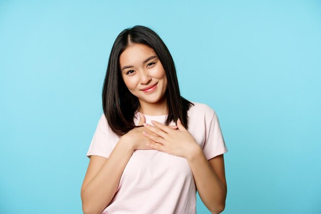 Smiling asian female model holds hands on heart, cherish smth, feeling care and warm love feelings, standing in t-shirt over blue background.