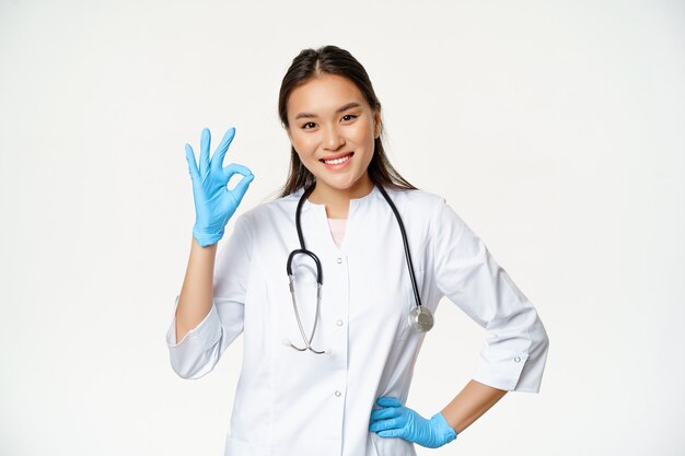 Smiling asian female physician, nurse in rubber gloves and medical uniform shows okay sign, approves and likes smth good, standing over white background