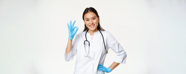 Free photo smiling asian female physician nurse in rubber gloves and medical uniform shows okay sign approves a