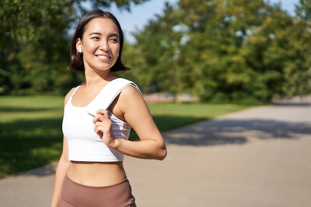 Free photo smiling asian fitness girl holding towel on shoulder workout in park sweating after training exercis