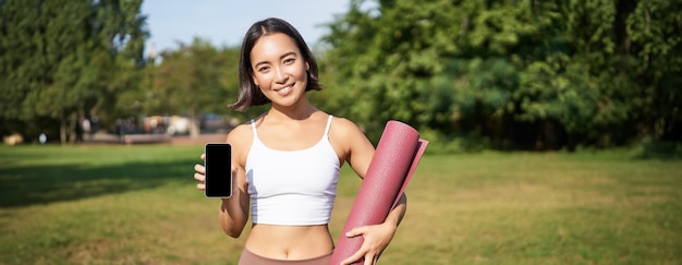 Free photo smiling asian fitness girl with rubber yoga mat shows her smartphone screen recommends workout