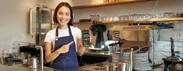 Foto gratuita una ragazza asiatica sorridente, barista, proprietaria di un caffè in grembiule, che mostra un lettore di carte di pagamento.