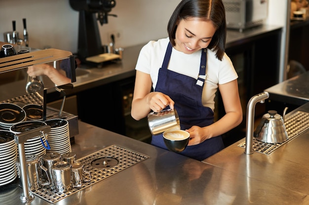 Free photo smiling asian girl barista cafe staff pouring steamed milk in coffee prepare cappuccino with latte a