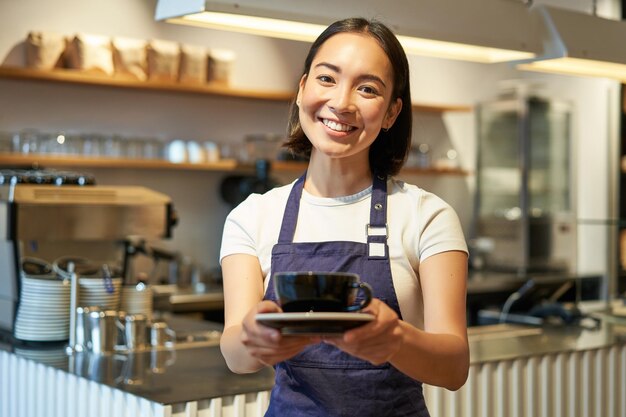 카운터 근처에 서있는 카페 유니폼 앞치마를 입고 커피를 들고 웃는 아시아 소녀 바리 스타