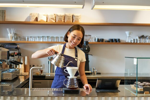 カウントの後ろに立ってコーヒーを醸造するやかんからお湯を注ぐ笑顔のアジアの女の子のバリスタ