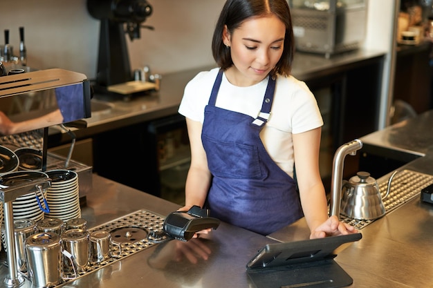 Free photo smiling asian girl barista working in cafe at counter processing orders using pos terminal receiving