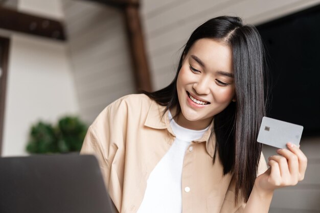Smiling asian girl buying online holding credit card in hand shopping on laptop from her home living...