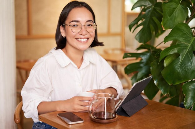 リモートでコーヒーを飲み、タブレットを使用して作業しているメガネの女性で笑顔のアジアの女の子