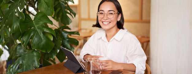 Free photo smiling asian girl in glasses woman working on remote drinking coffee and using tablet