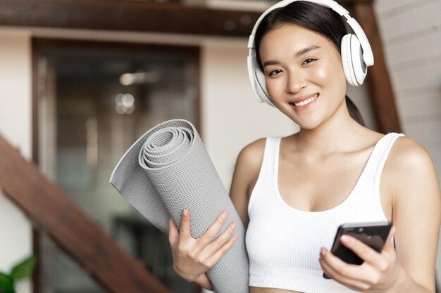自宅で音楽を聴いているヘッドフォンで笑顔のアジアの女の子とモバイルpを保持しているフロアマットでトレーニング...