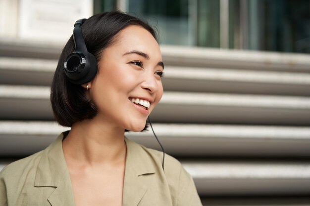 Smiling asian girl laughing listening music in headphones sitting outdoors uni student enjoying free