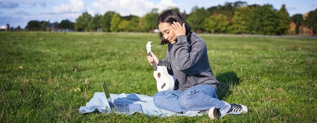 Free photo smiling asian girl learns how to play ukulele via laptop online video tutorials sitting on grass in