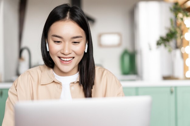 Улыбающаяся азиатская девушка слушает, смотрит на ноутбук, глядя на компьютер во время онлайн-вебинара, концепция ...