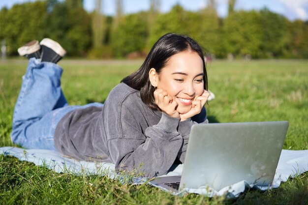 Free photo smiling asian girl lying in park on grass watching movie or video on laptop looking at screen with i