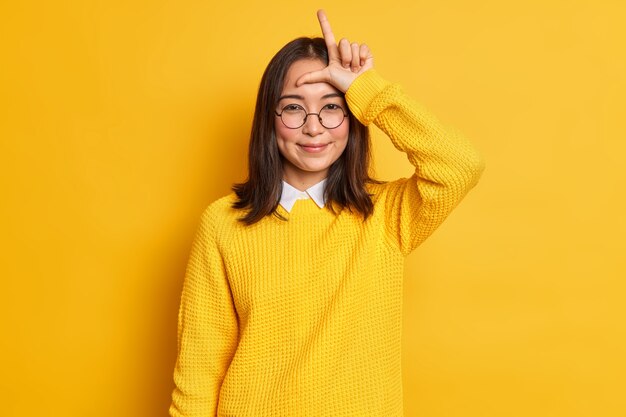 smiling Asian girl makes loser gesture over forehead looks with satisfaction says friend you lost shape L letter with fingers makes fun of team wears round glasses and sweater