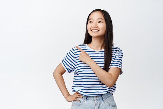 Smiling asian girl pointing finger and looking left standing in tshirt and jeans on white.