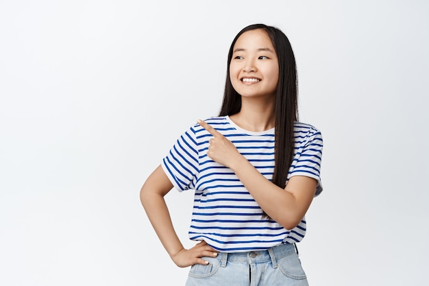 웃는 아시아 소녀가 손가락을 가리키고 흰색 티셔츠와 청바지를 입고 왼쪽을 바라보고 있습니다.