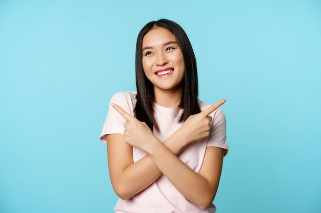 Smiling asian girl pointing sideways, showing two choices and laughing, picking between variants, blue background.