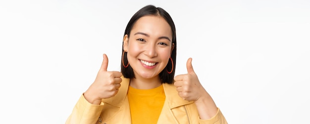 Smiling asian girl showing thumbs up looking pleased approve smth standing over white background Copy space