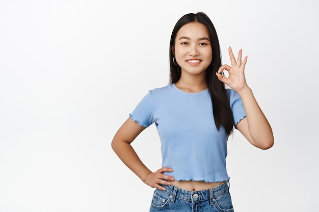 Smiling asian girl shows okay gesture confirm something say yes making Ok zero sign standing over white background