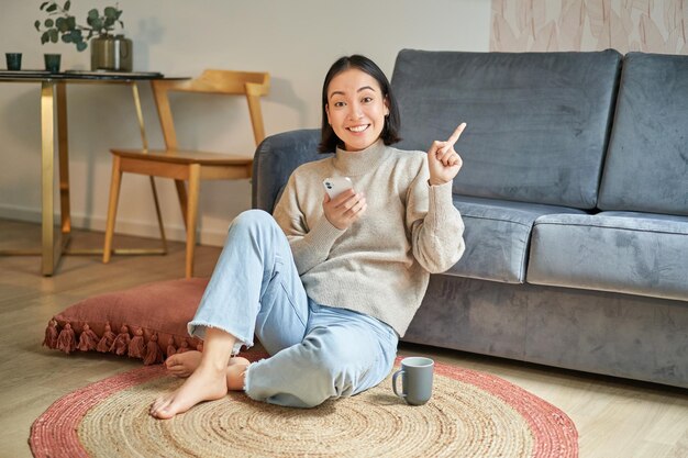 smiling asian girl sits on floor in stylish living room pointing finger at advertisement showing promo banner holding mobile phone in hand