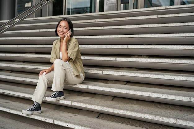 Бесплатное фото Улыбающаяся азиатка сидит на лестнице здания и разговаривает по мобильному телефону, расслабляясь во время телефонного разговора
