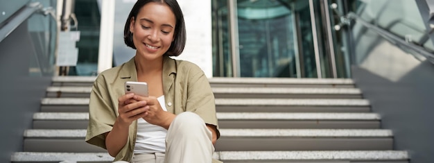 Free photo smiling asian girl sits on stairs near building entrance using mobile phone app happy young woman