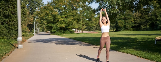 Foto gratuita ragazza asiatica sorridente che si allunga dopo un buon allenamento nel parco ascoltando musica con le cuffie wireless