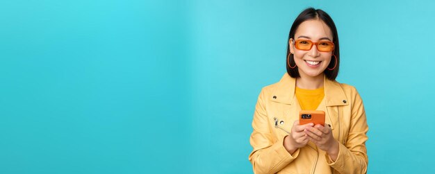 Smiling asian girl in sunglasses using smartphone app holding mobile phone standing over blue background