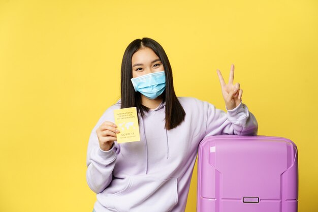얼굴 마스크를 쓴 웃는 아시아 소녀 관광객, 여행 가방을 들고 서서 여행자를 위한 코비드 국제 예방 접종 증명서와 평화 표지, 노란색 배경을 보여줍니다.