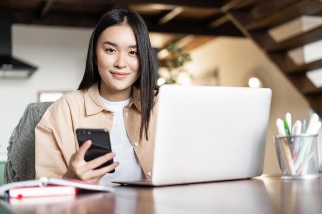 Smiling asian girl uses computer at home holds smartphone and looks happy at camera busy woman worki...