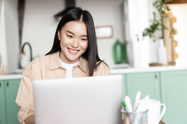 Smiling asian girl using laptop working from home on computer pc sitting in kitchen and studying doi...