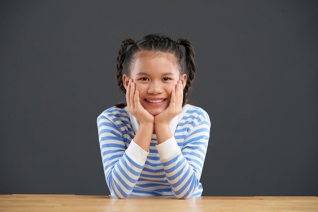 Free photo smiling asian girl with braids sitting at table with hands on cheeks