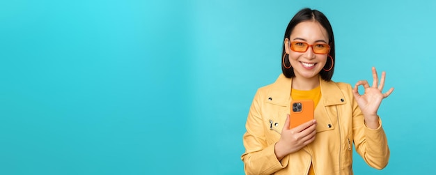 파란색 배경 복사 공간 위에 승인 표시를 하는 스마트폰을 들고 웃고 있는 아시아 소녀