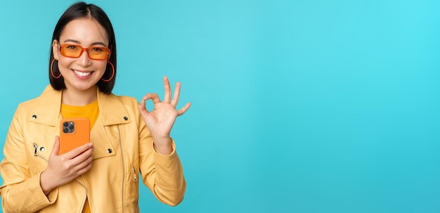 Free photo smiling asian girl with smartphone shwoing okay ok sign in approval standing over blue background